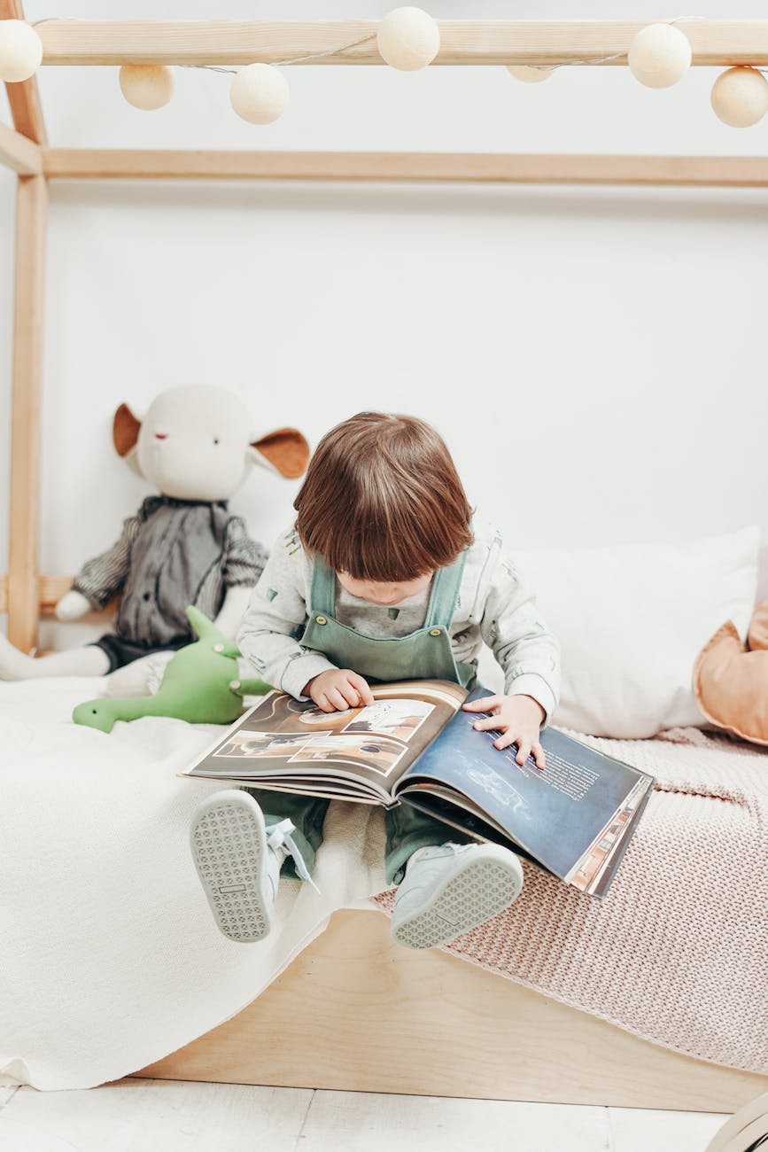 child in white long sleeve top and dungaree trousers sitting on bed reading book